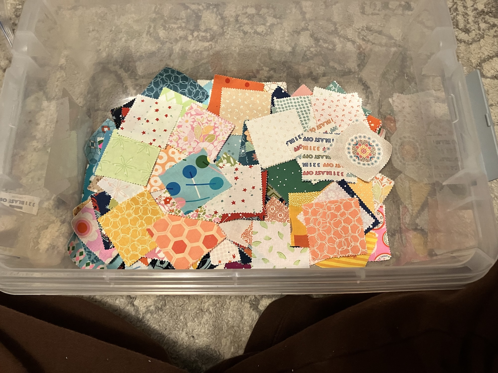 A clear plastic bin holding the scattered aforementioned 2.5 inch fabric squares. They are brightly colored with many different patterns and writings