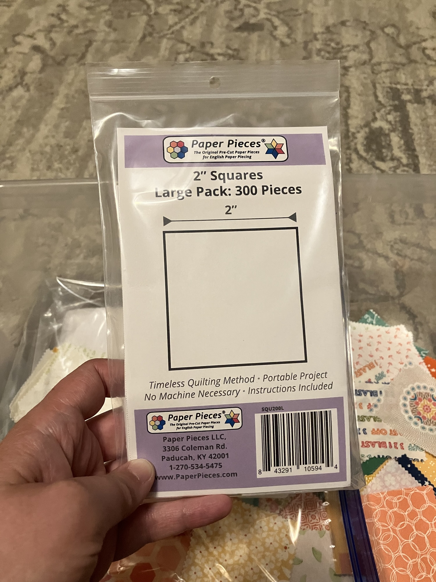 A white woman’s hand holding a plastic bag containing 300 pieces of 2 inch cardboard squares. The company is Paper Pieces LLC and their website is www.paperpieces.com