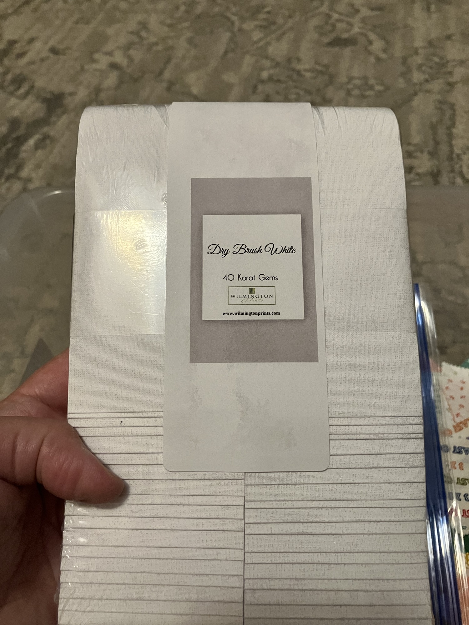 A white woman’s hand holding a package of white strips of fabric that are 2.5 inches wide. The label reads Dry Brush White, 40 Karat Gems, and the maker is Wilmington Prints. Their website is listed as www.wilmingtonprints.com