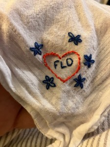 The corner of a white handkerchief with simple embroidery of a pink heart with blue initials and flowers 